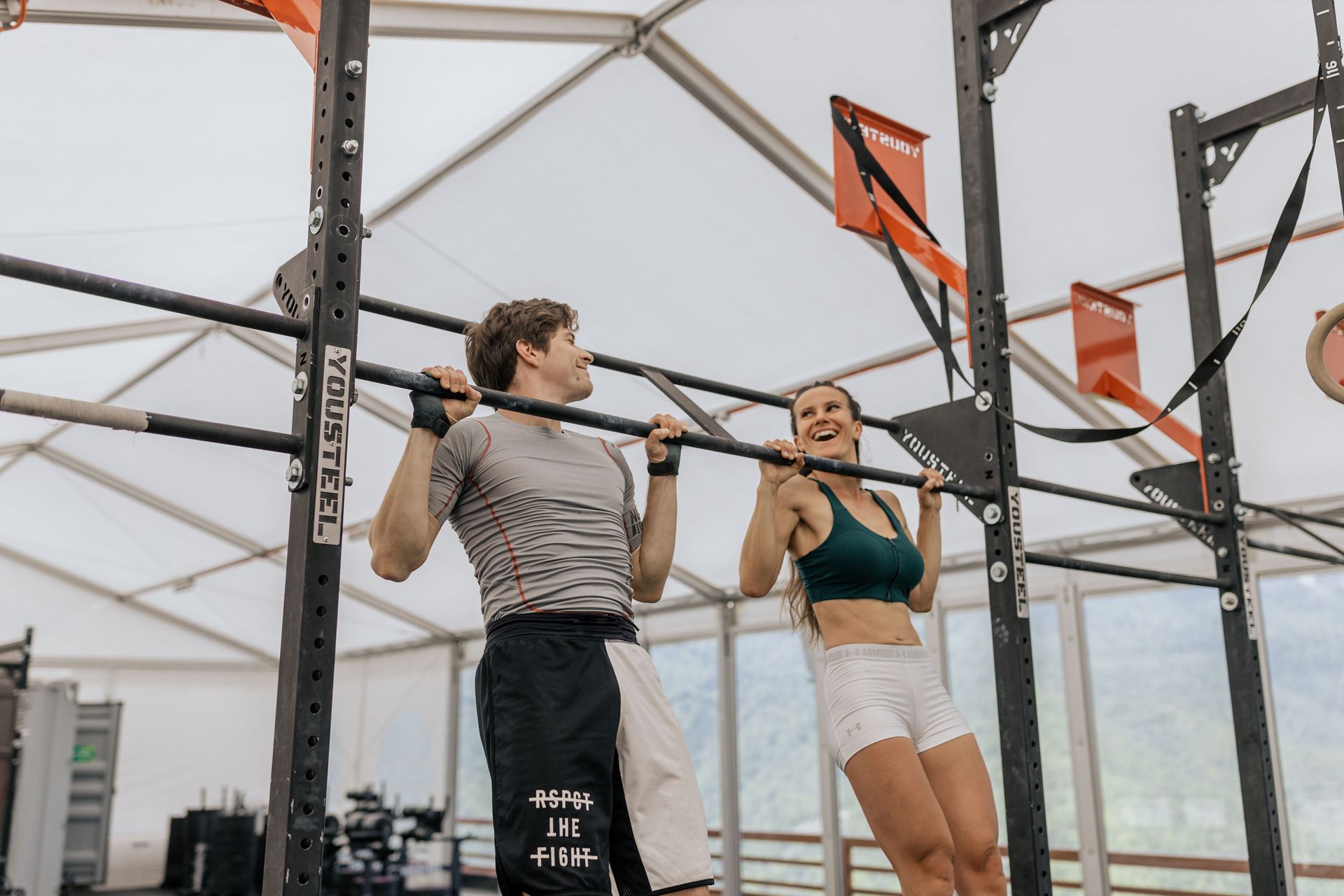 Two athletes doing pull-ups in a gym, with black rigging and orange flags overhead.