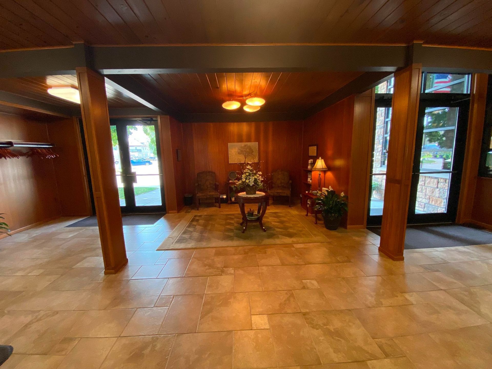 Lobby with wood paneling, tile floor, chairs, and table with flowers. Two doors lead outside, lit by ceiling lights.