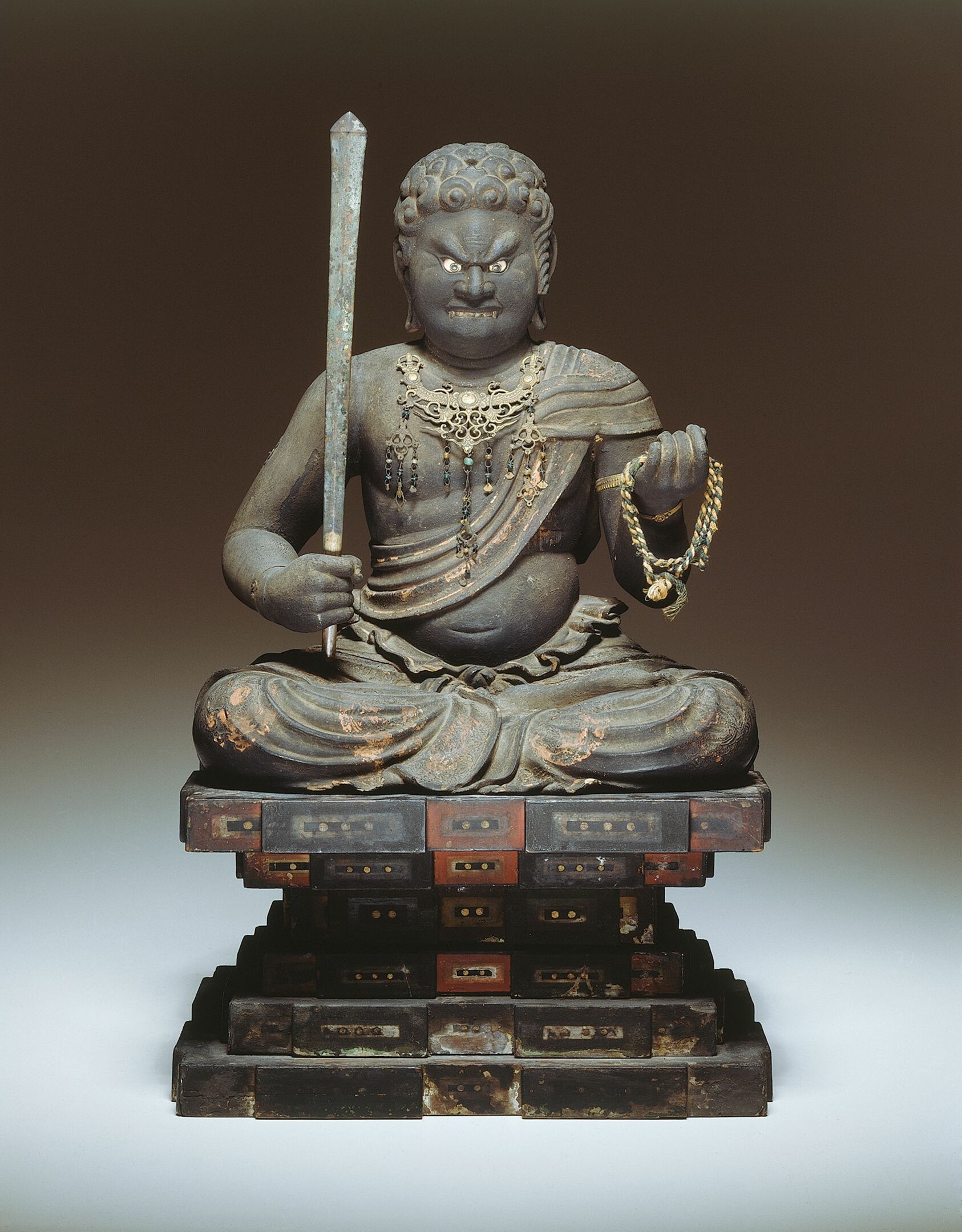 Seated Buddhist deity with sword. Dark, carved figure on layered pedestal.
