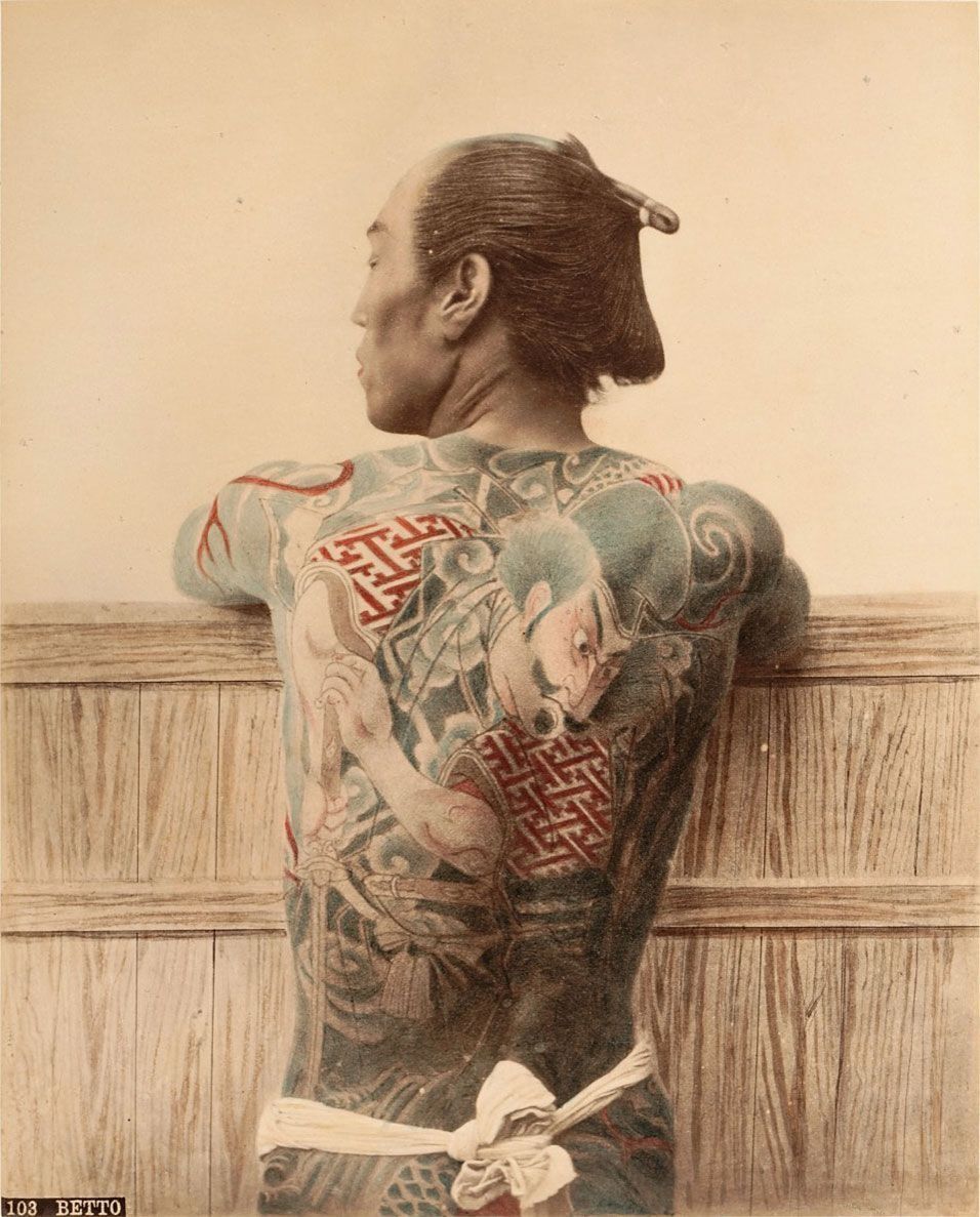 Man with full-body tattoo stands near a wooden structure, back facing the viewer. The tattoo includes red, blue, and green ink.