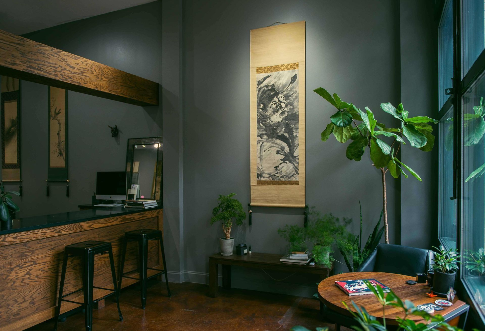 Cozy tattoo studio corner with dark gray walls, artwork, plants, and a wooden bar with stools.