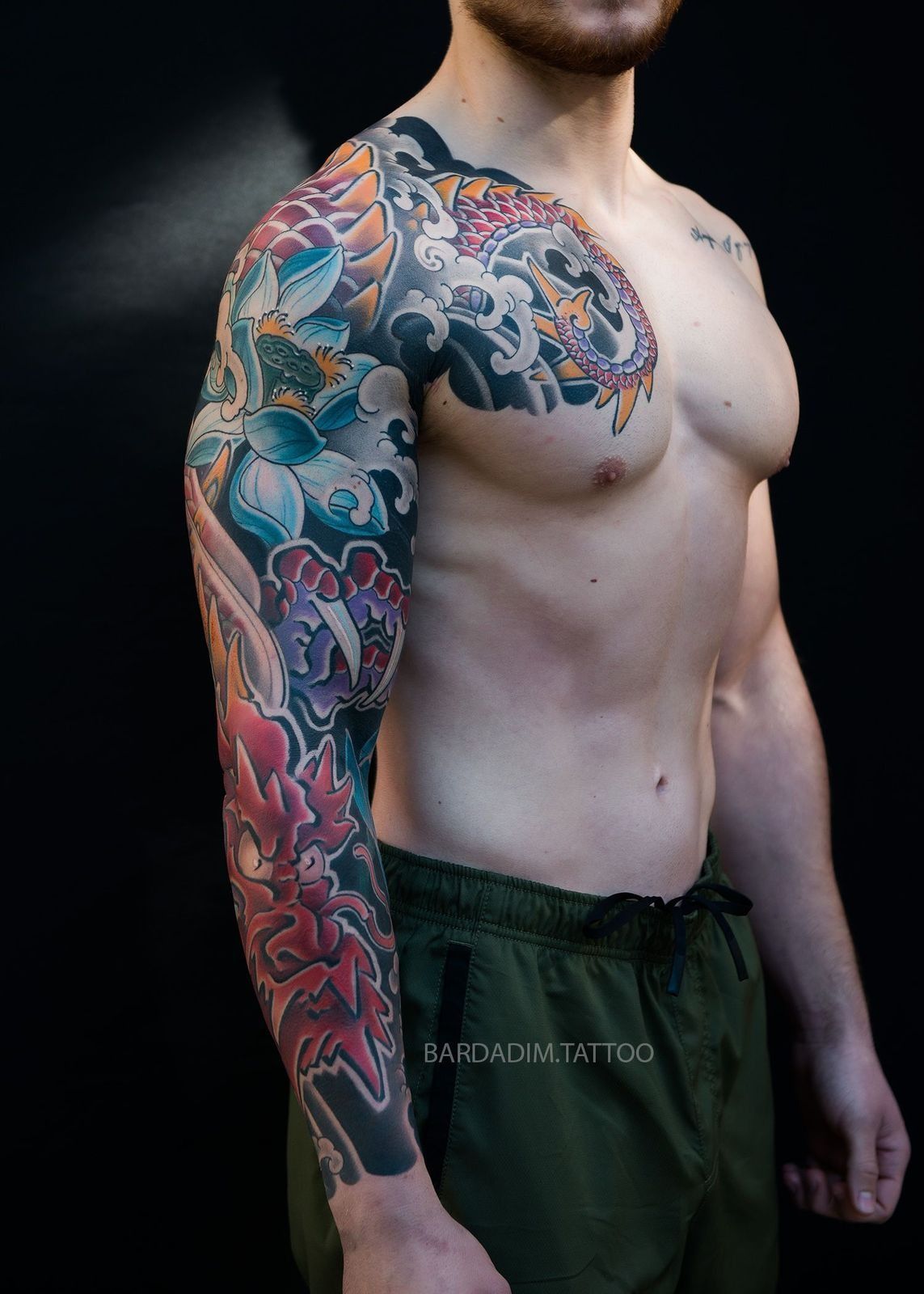 Man with full sleeve tattoo of colorful flowers, waves, and a dragon, against a black background.