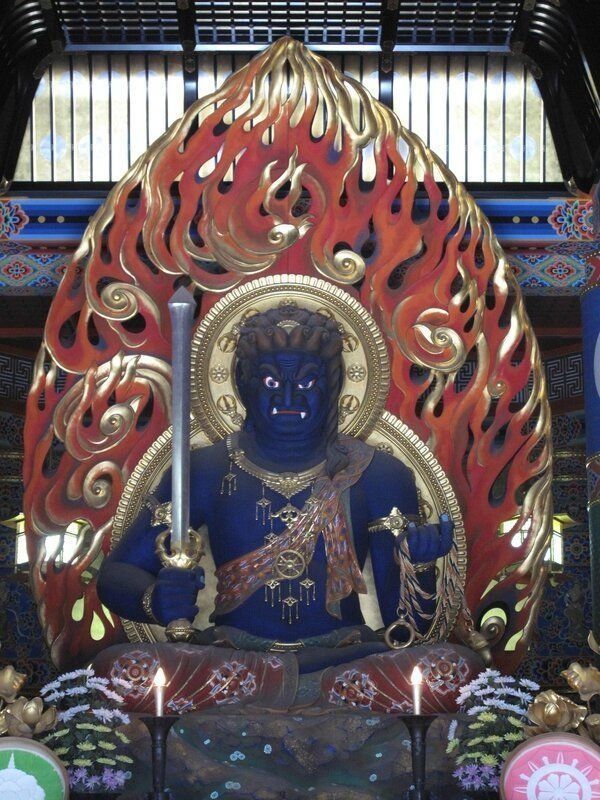 Blue-skinned Buddhist deity with fiery halo, holding a sword and jewel, seated in a temple.