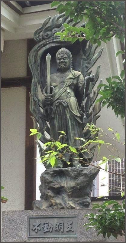 Statue of Fudo Myoo, a wrathful deity, holding sword, surrounded by flames, outside a building.