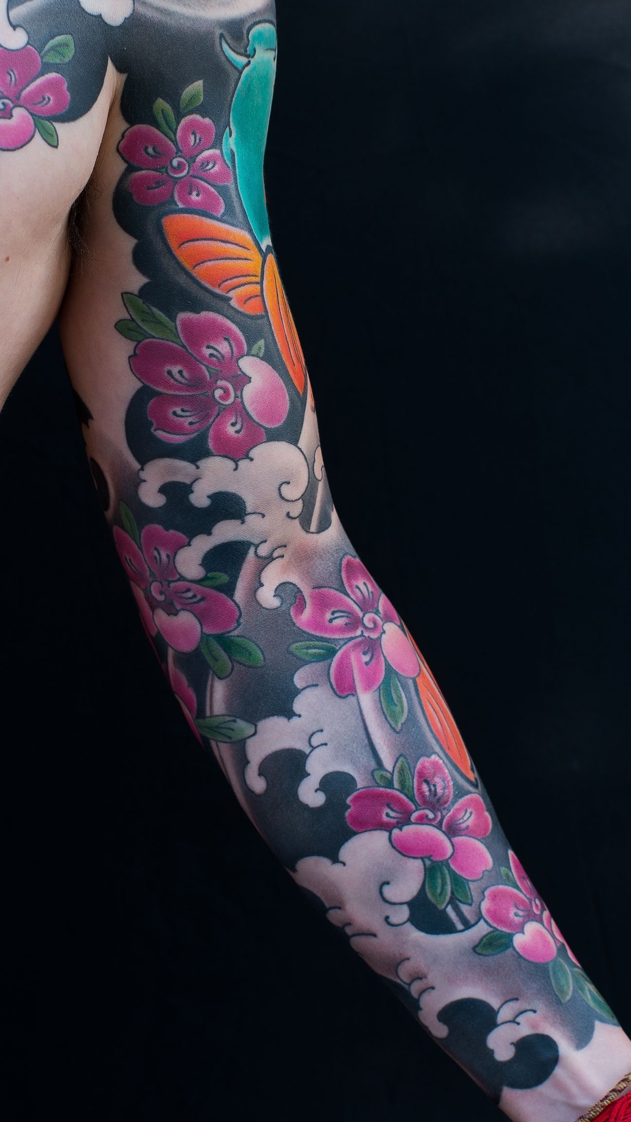 Tattoo sleeve depicting a koi fish, cherry blossoms, and water waves, in vibrant colors on a dark background.