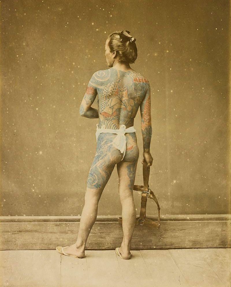 A man with a lot of tattoos on his body is standing in front of a wall.