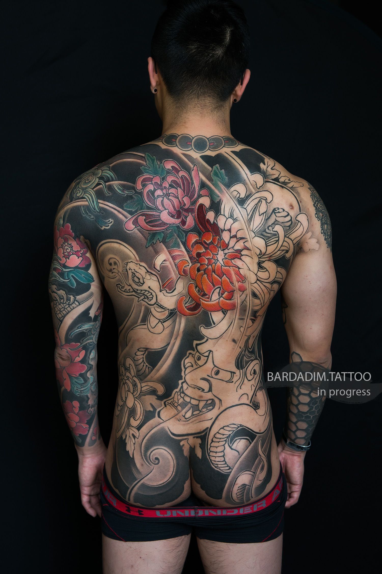 Man's back covered in elaborate, colorful Japanese-style tattoo against a black backdrop.