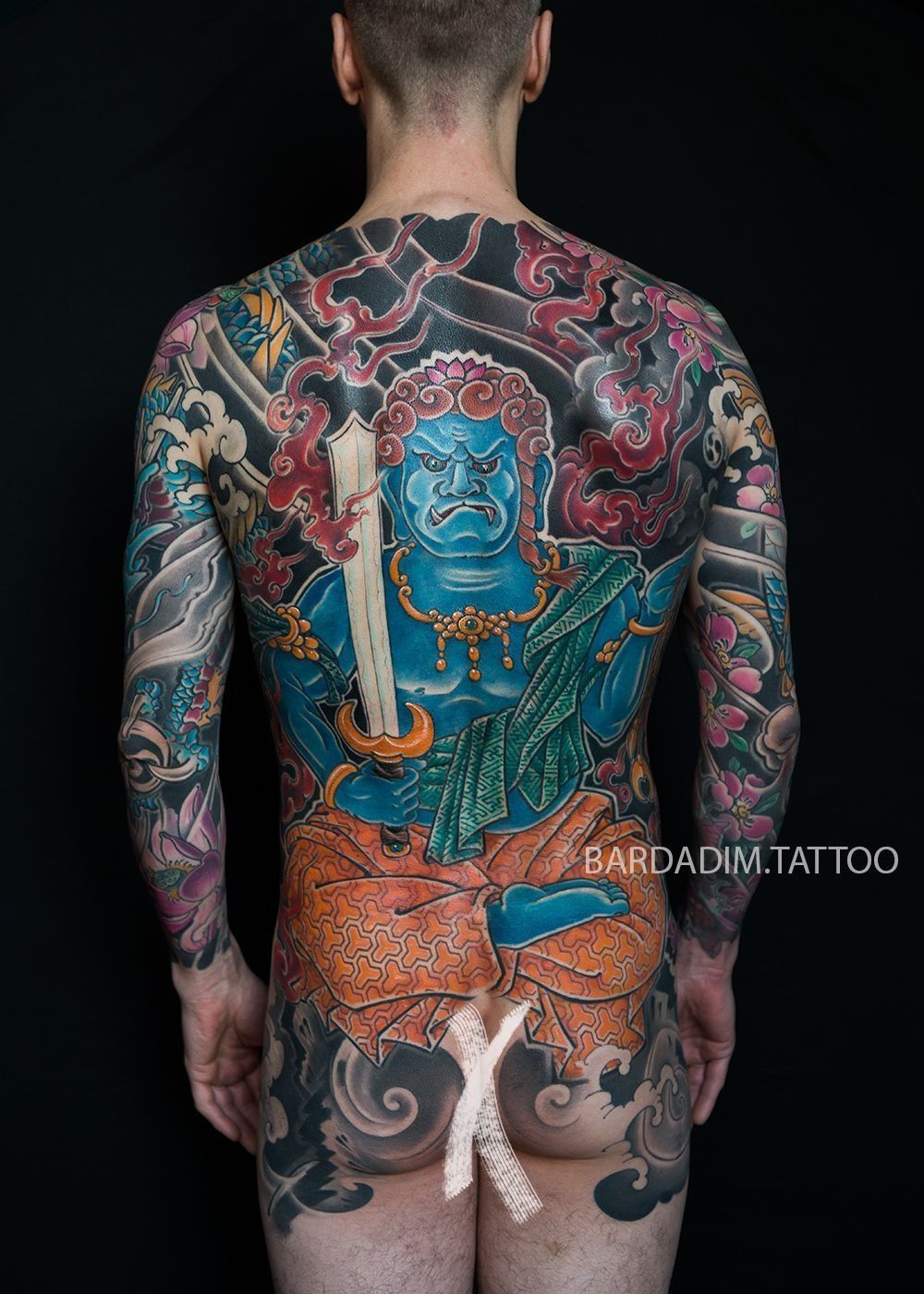 Man's back covered in elaborate, colorful Japanese-style tattoo featuring a blue deity holding a sword.