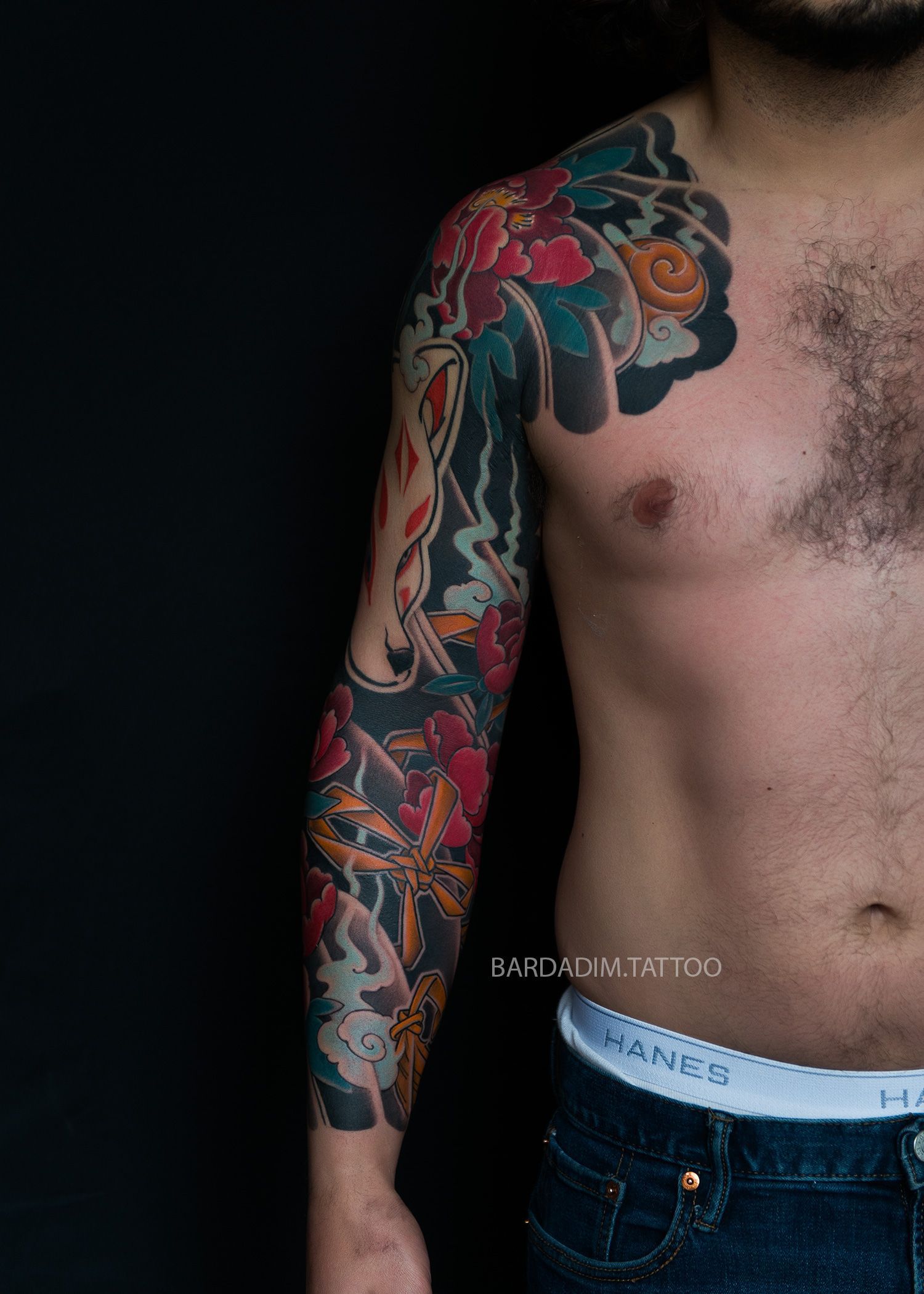 Man with elaborate, colorful arm sleeve tattoo. Dark background, of Bardadim Tattoo