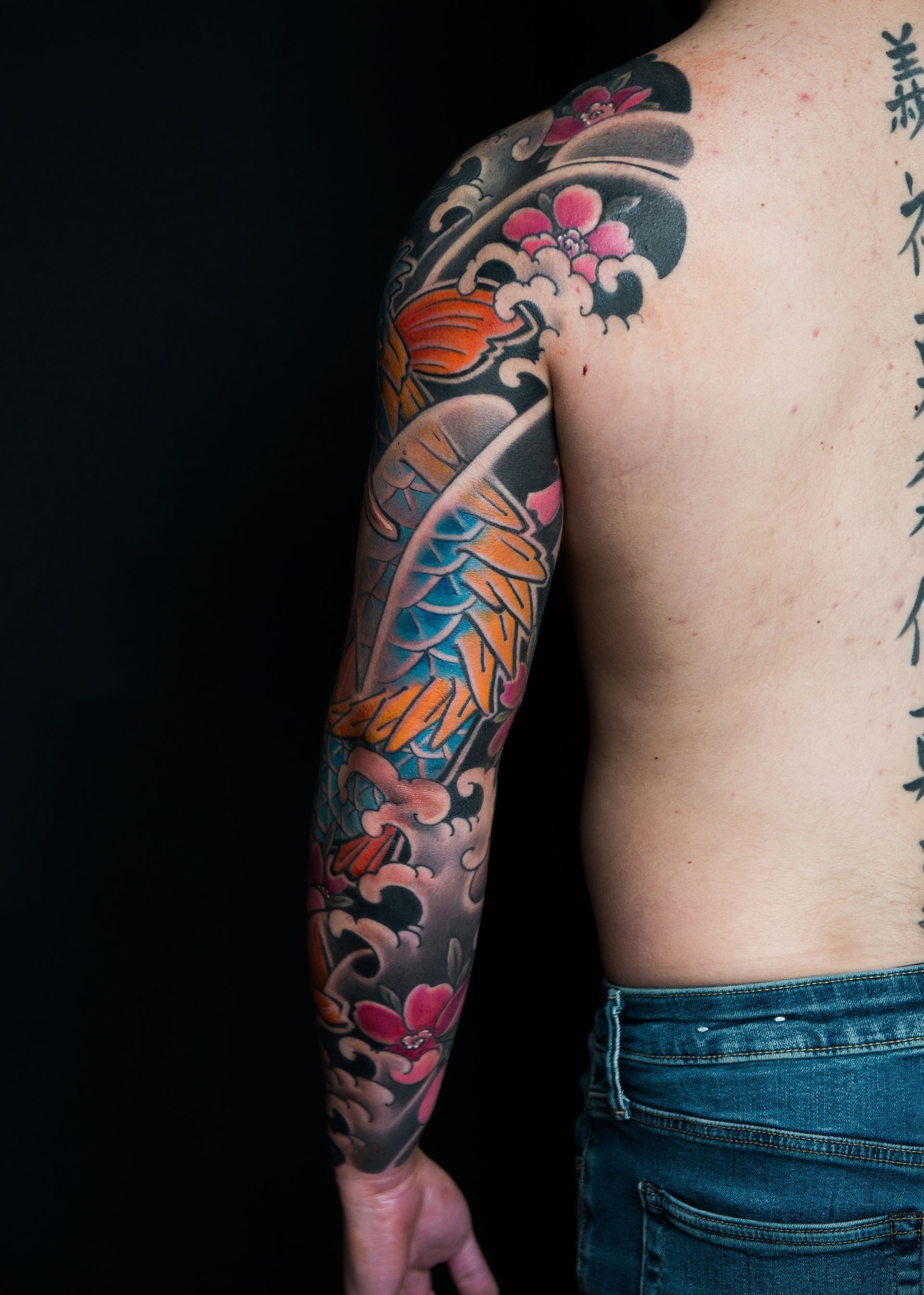 Man's arm covered in colorful koi-dragon and  cherry blossoms tattoo, dark background.