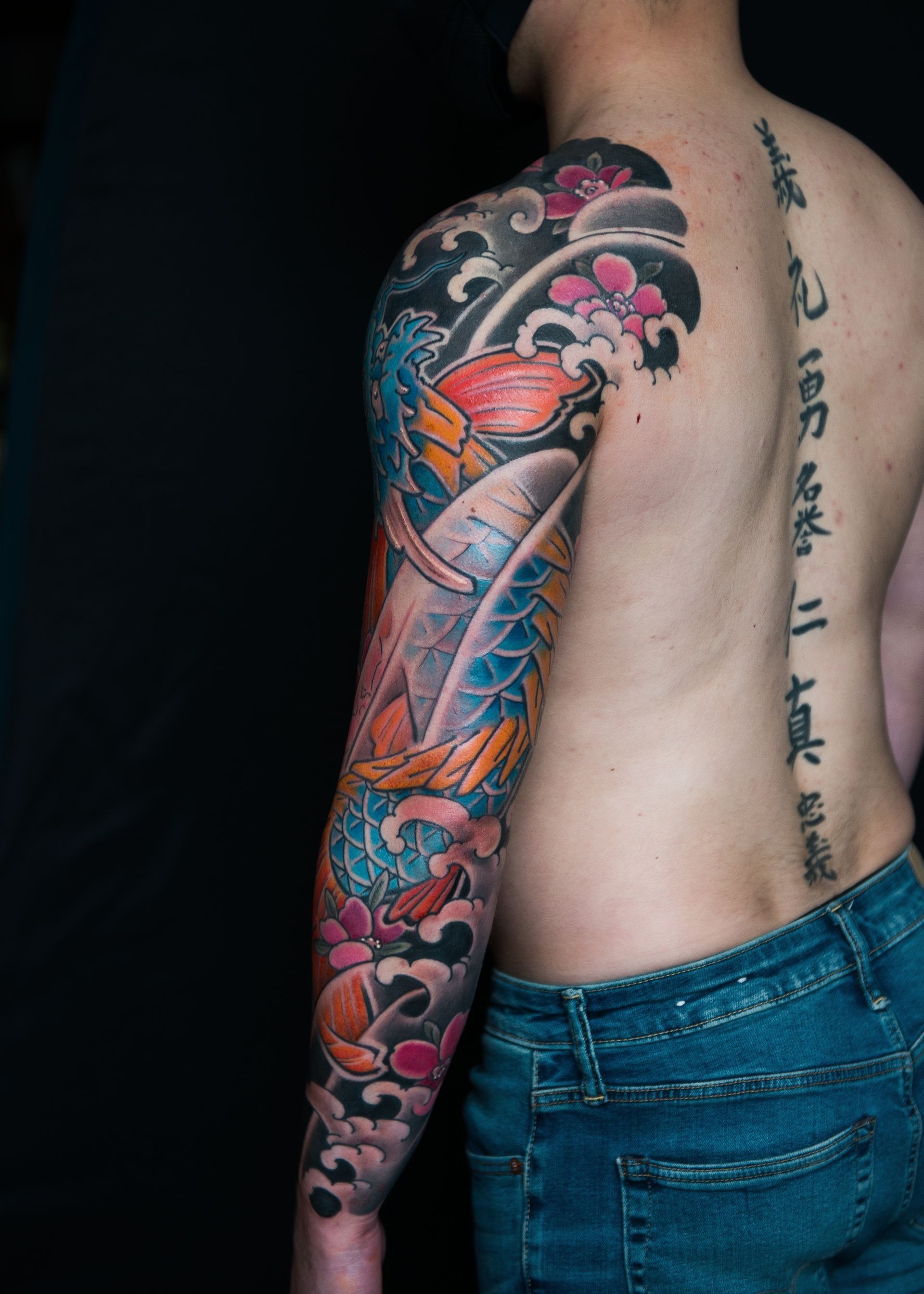 Man with a full sleeve tattoo of a koi-dragon, sakura flowers, and waves; black background.