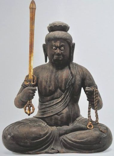 Statue of a seated figure of Acala holding a sword and prayer beads. Dark brown wood, fierce expression.