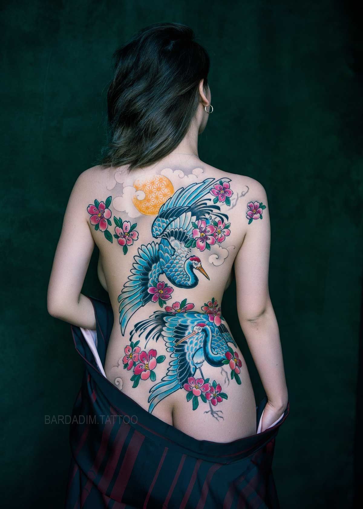 Woman with crane and flower back tattoo, dark background.