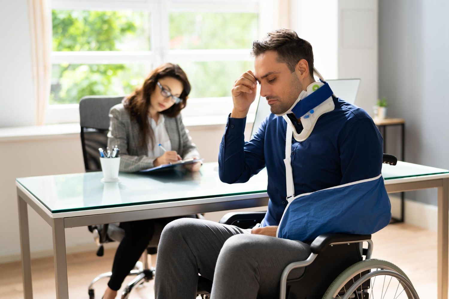 Man in wheelchair, arm in sling, neck brace, consulting with a woman, possibly a lawyer, indoors at desk.