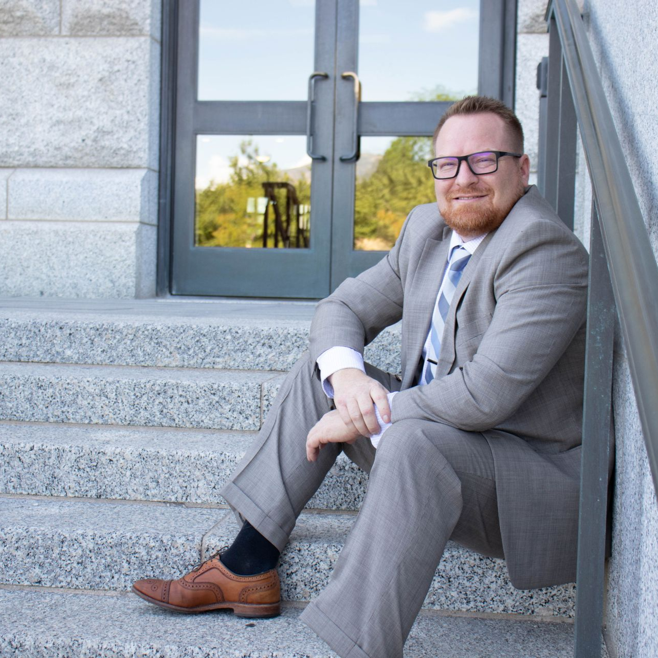 Man in suit sits on stone steps, smiling. Glasses, red beard, building background.