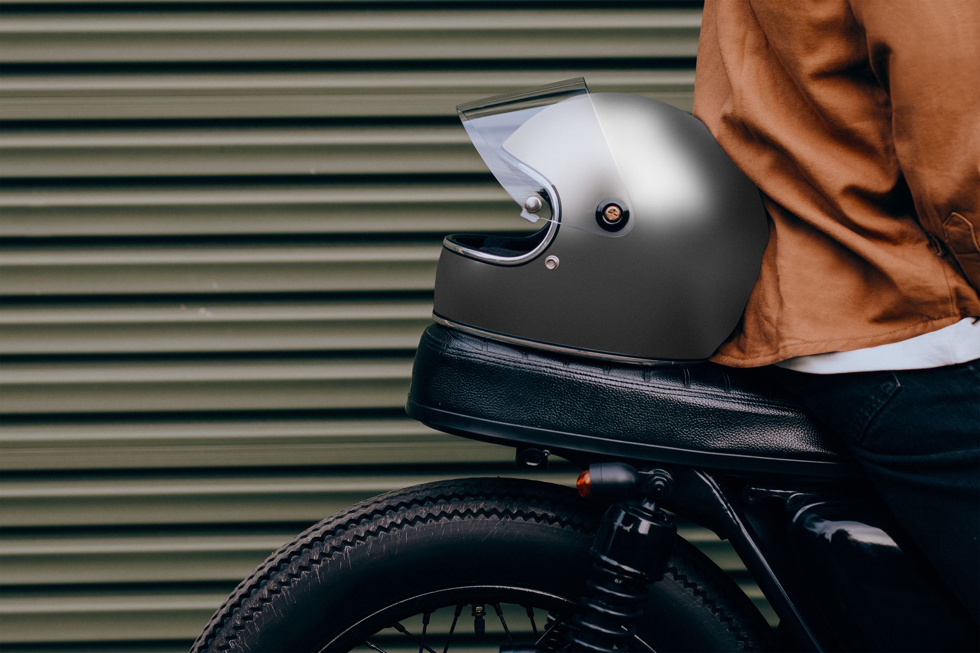 Motorcycle helmet on a black seat; person wearing a brown jacket, green corrugated background.