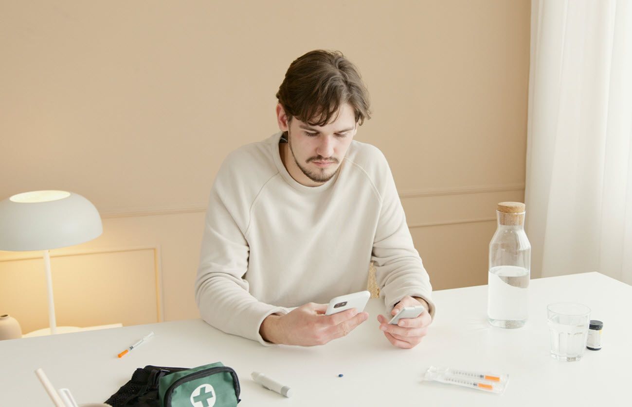 Man checks phone while preparing diabetes supplies on a white table. Light beige wall in background.