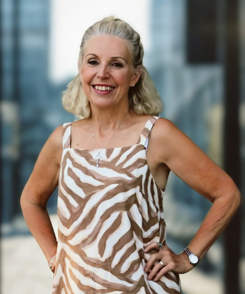 Woman with blonde hair wearing a beige and white zebra-print dress, smiling with hands on hips, standing outside.