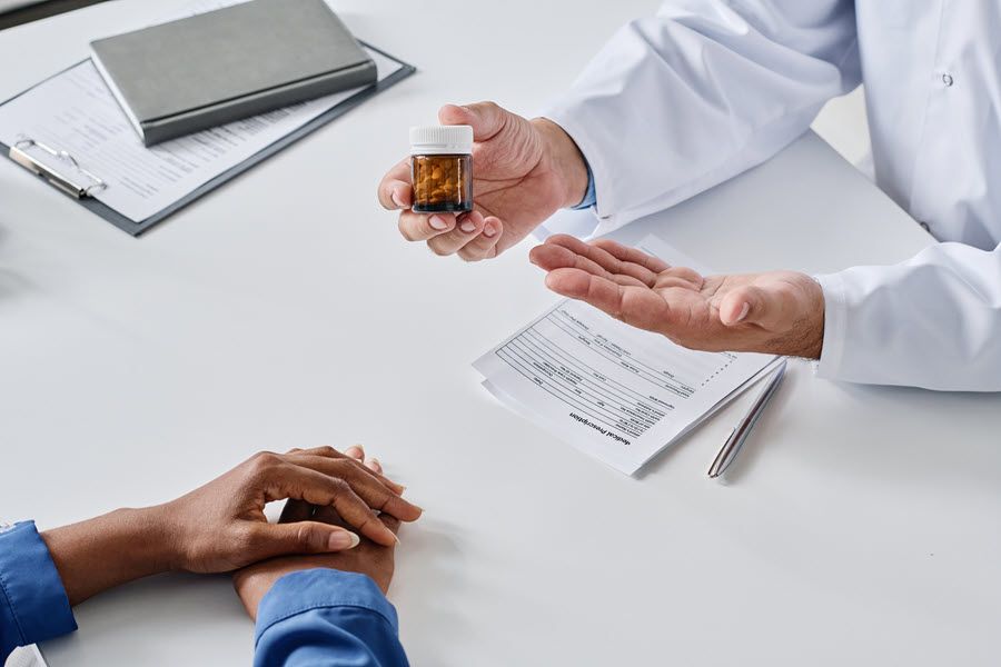 Doctor holding pill bottle, gesturing toward patient at a table with prescription paperwork.