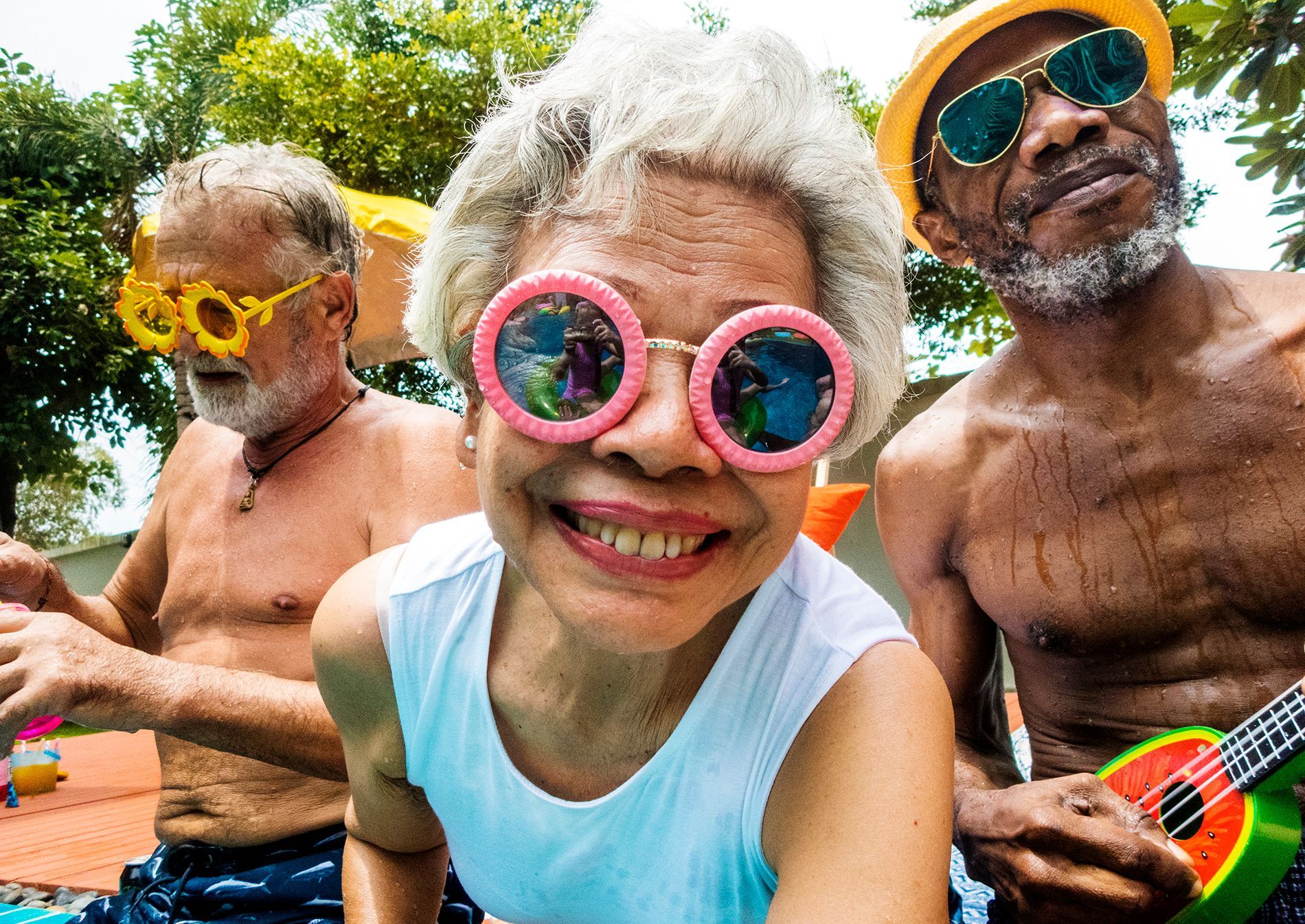 Three older adults having fun outdoors, wearing sunglasses. One holds a toy ukulele.