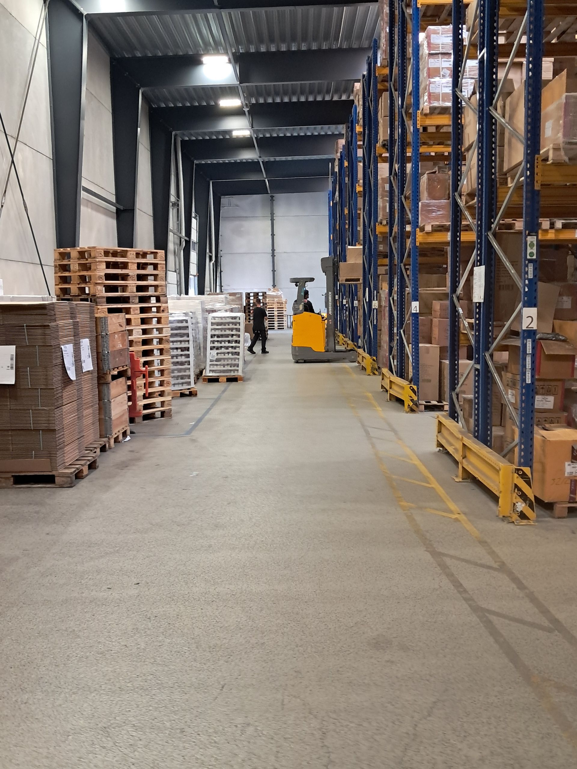 A warehouse aisle with tall storage racks, pallets on the left, and a forklift with a driver further down the path.