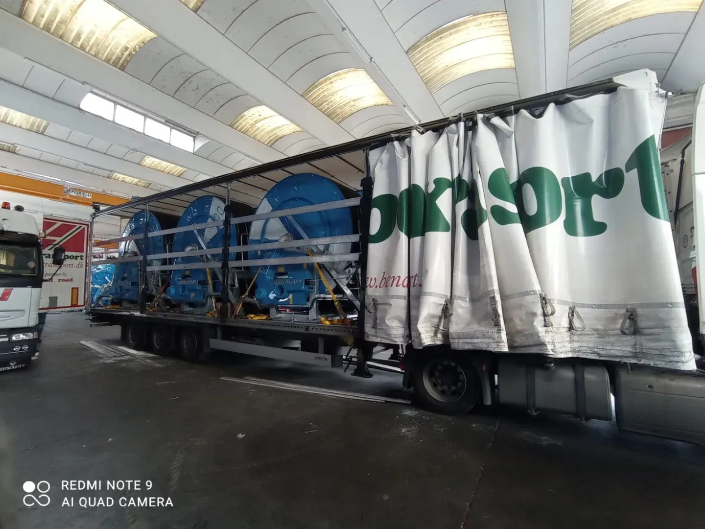 En semitrailer læsset med blåt industrimaskineri inde i et lager. Traileren har hvide sidegardiner med grøn tekst.