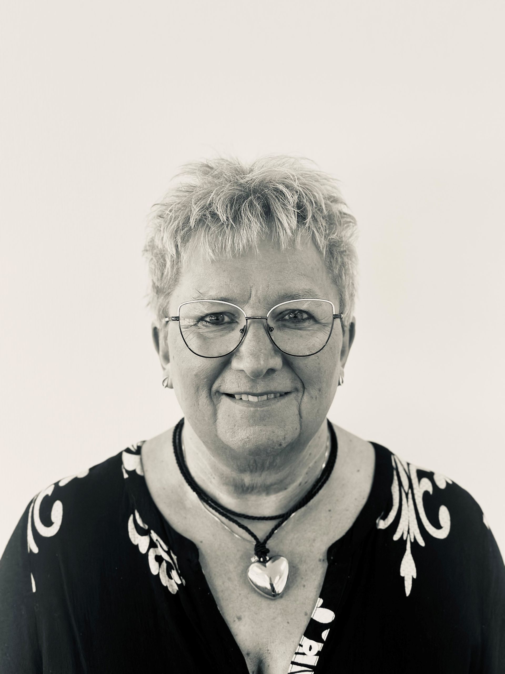 Smiling person with short hair, glasses, and a necklace, wearing a patterned top against a plain background