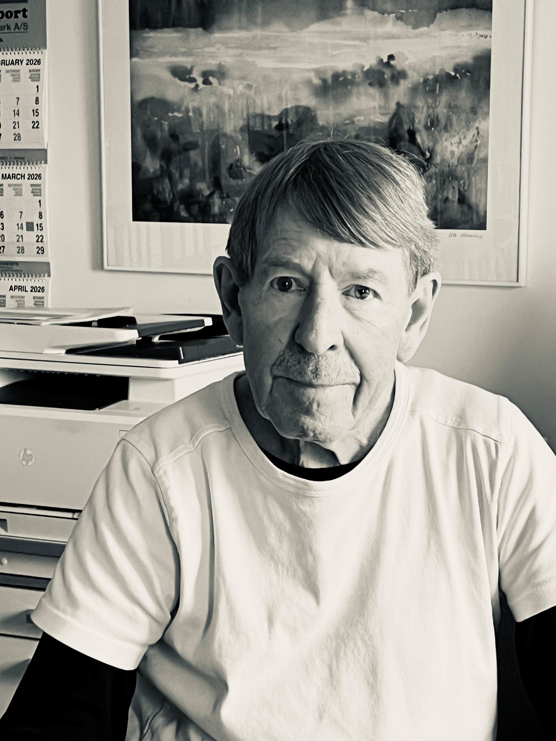 Person seated indoors in a white shirt, looking at the camera, with framed artwork in the background