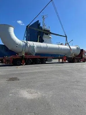 Large white industrial pipe section being hoisted by a crane on a flatbed trailer under a blue sky.
