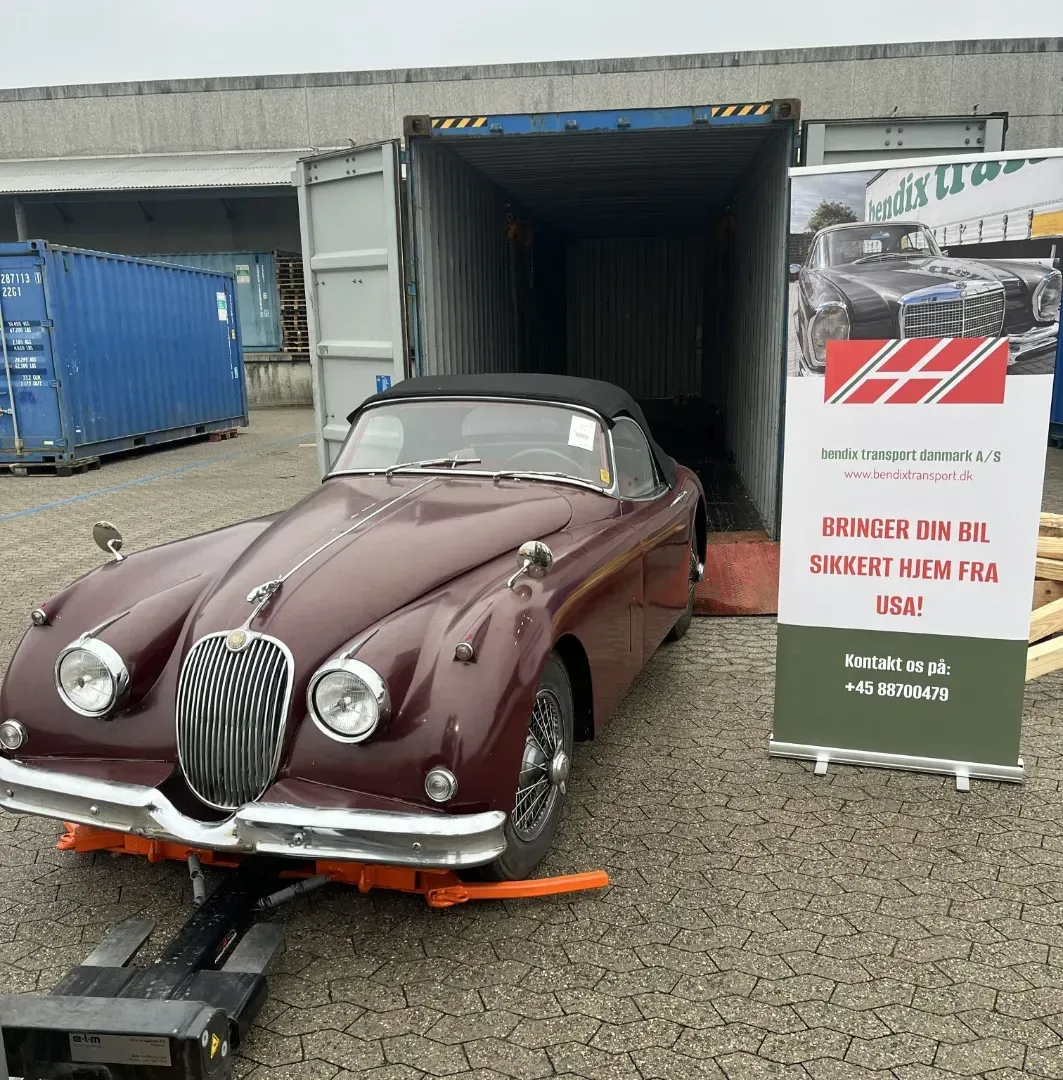 Vintage maroon convertible loaded into a shipping container beside a sign at an industrial site