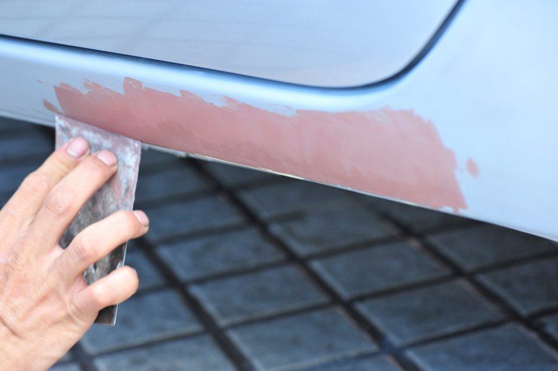 Persona che carteggia lo stucco su un pannello azzurro di un'auto all'aperto.
