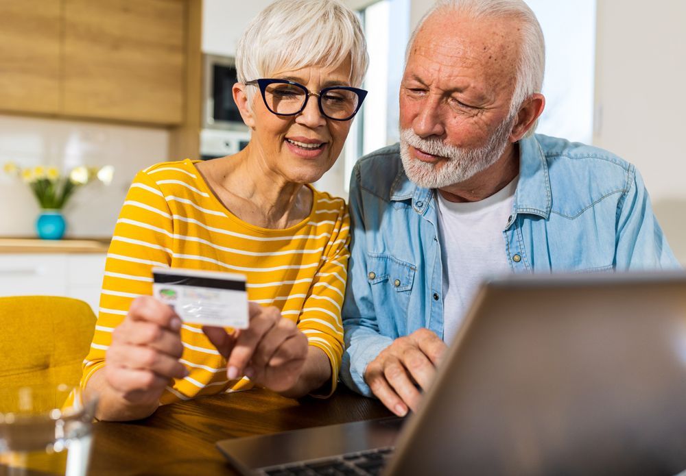 An elderly couple is sitting at a table holding a credit card and a laptop.