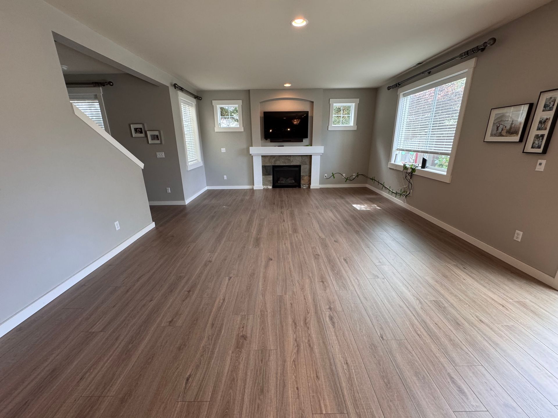 Empty living room with wood-look flooring, fireplace with TV, and windows.