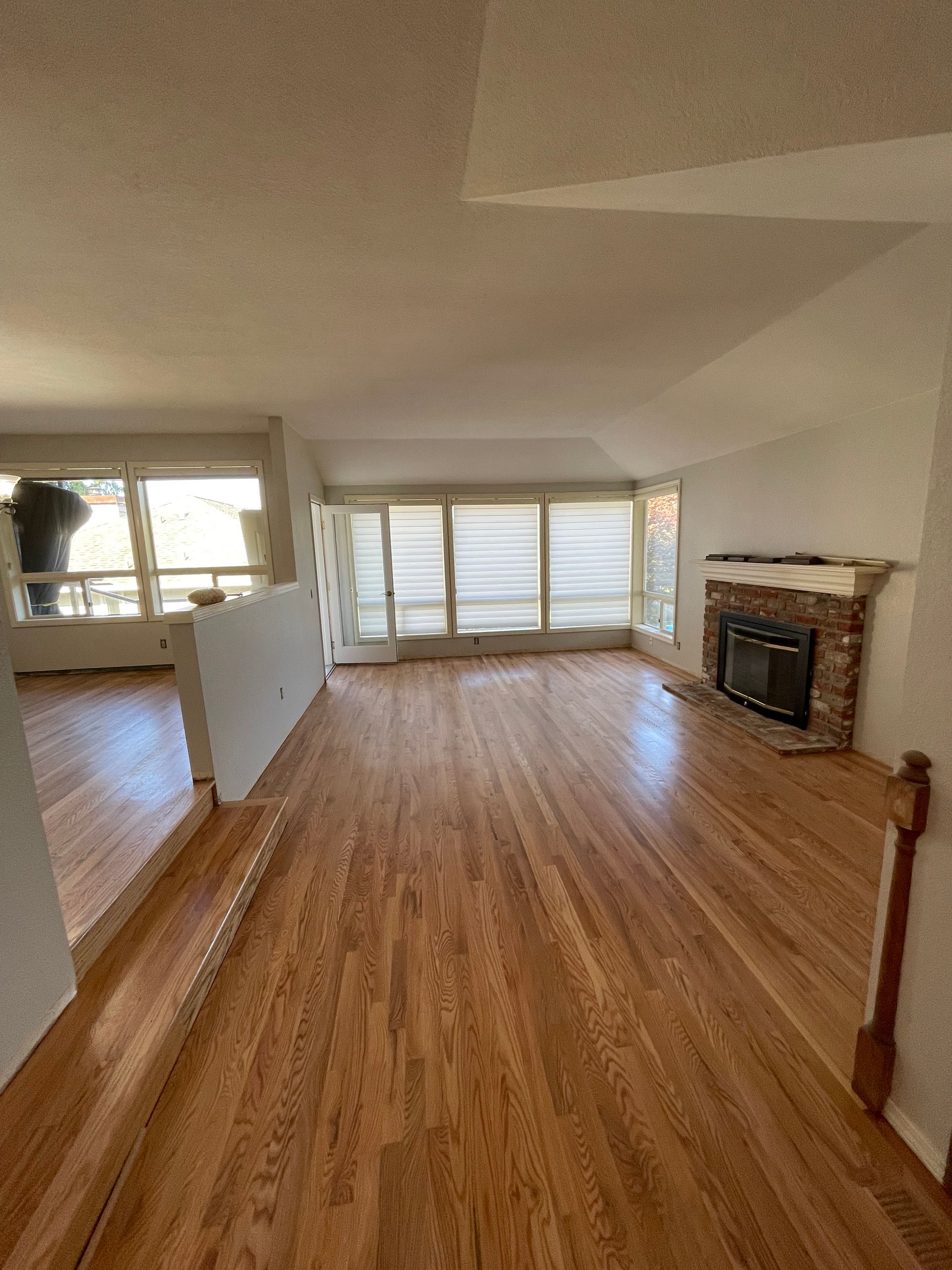 Wooden-floored living room with fireplace and large windows. Sunlight streams in.
