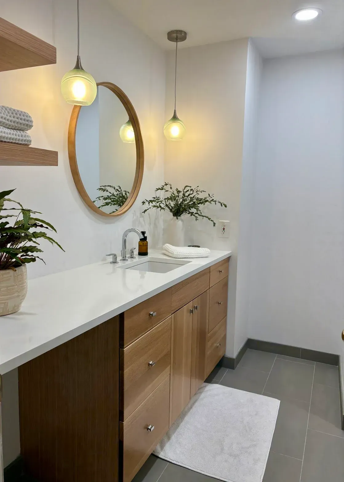 Modern bathroom with wooden vanity, round mirror, and two pendant lights. White countertop, grey floor, plants.