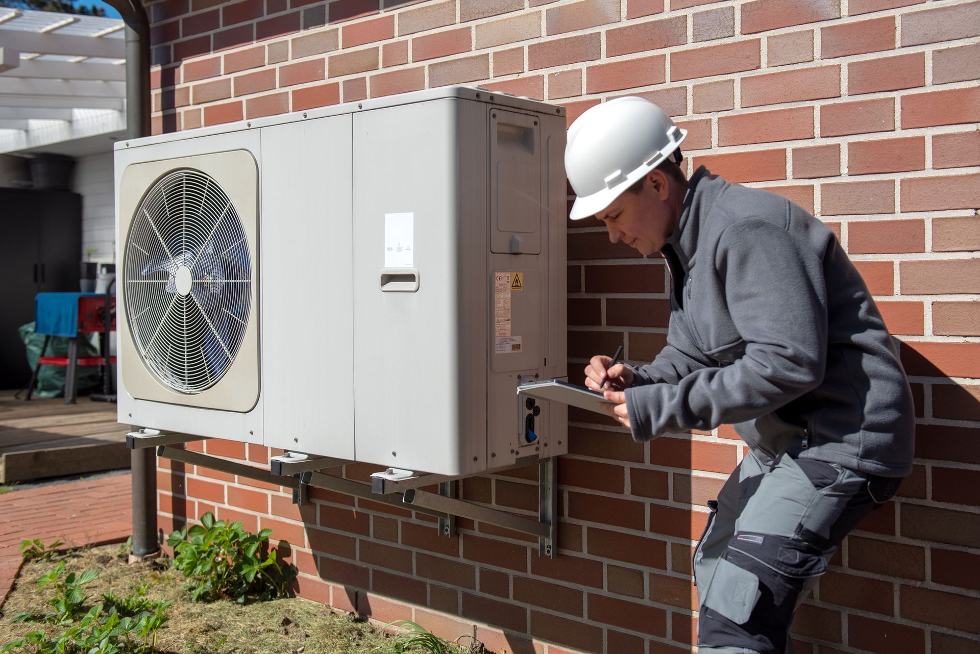Technician inspecting HVAC unit mounted on brick wall outdoors in daylight.