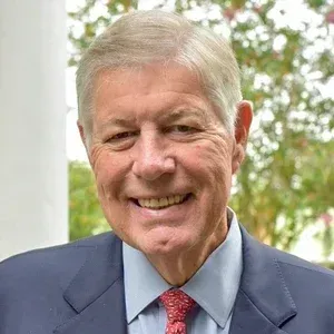 Smiling man in a blue suit and red tie outdoors with blurred greenery behind him