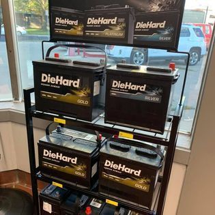 DieHard car batteries on a black display rack. Gold and silver batteries are visible. Store interior with a window in background.