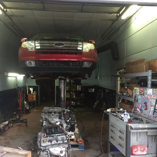 Red car raised on a lift inside a cluttered auto repair shop, engine parts on the floor.