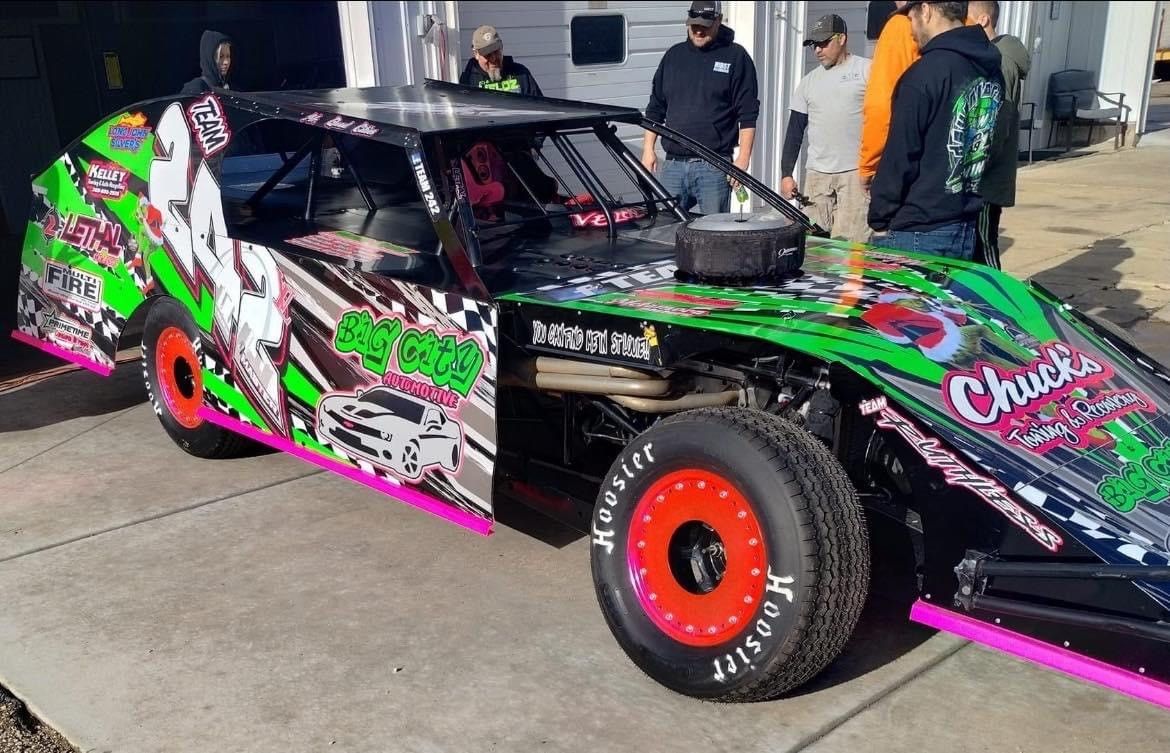 Modified race car with black, green, and pink graphics, surrounded by a group of people.