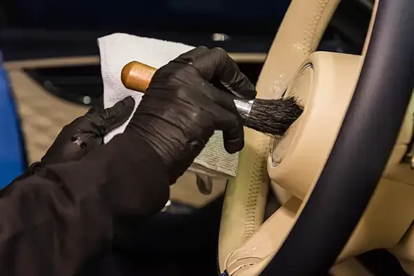 Gloved hand cleaning a beige car interior with a brush and cloth
