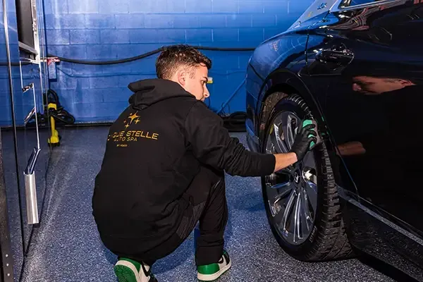 Detailer crouches beside a black car, cleaning the front wheel in a blue garage.