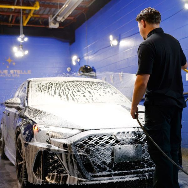 Car being washed with foam in a blue auto detailing bay, worker standing beside it.