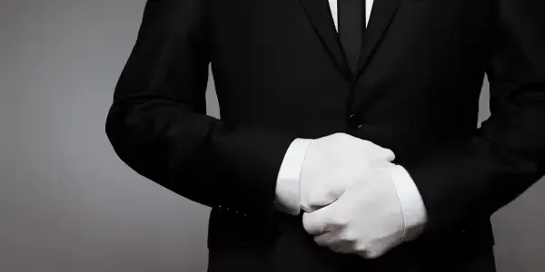 Person in a black suit with white gloves clasping their hands against a gray background