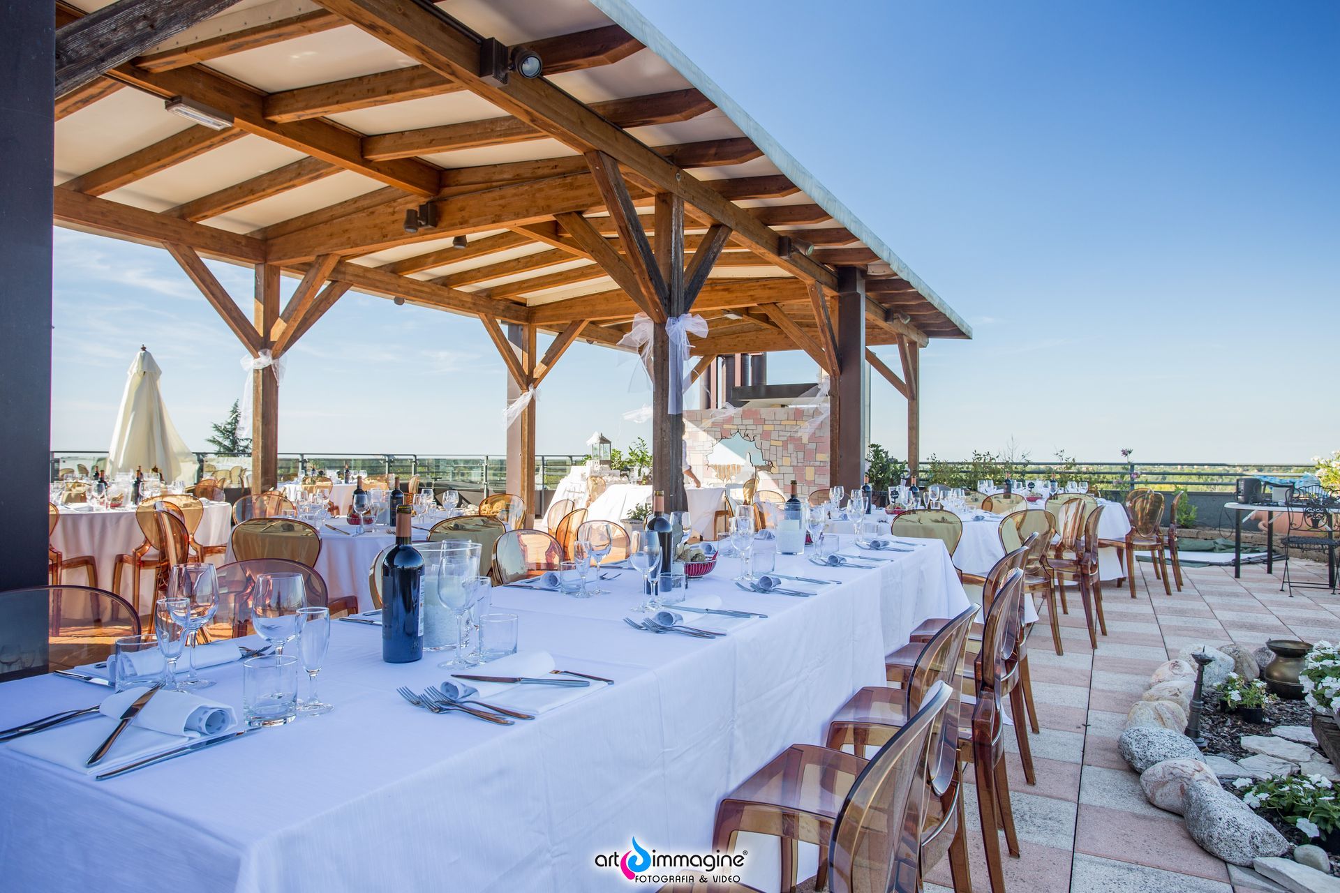 Tavolo da ricevimento all'aperto, lunga tovaglia bianca, sotto un pergolato in legno, cielo azzurro sullo sfondo.