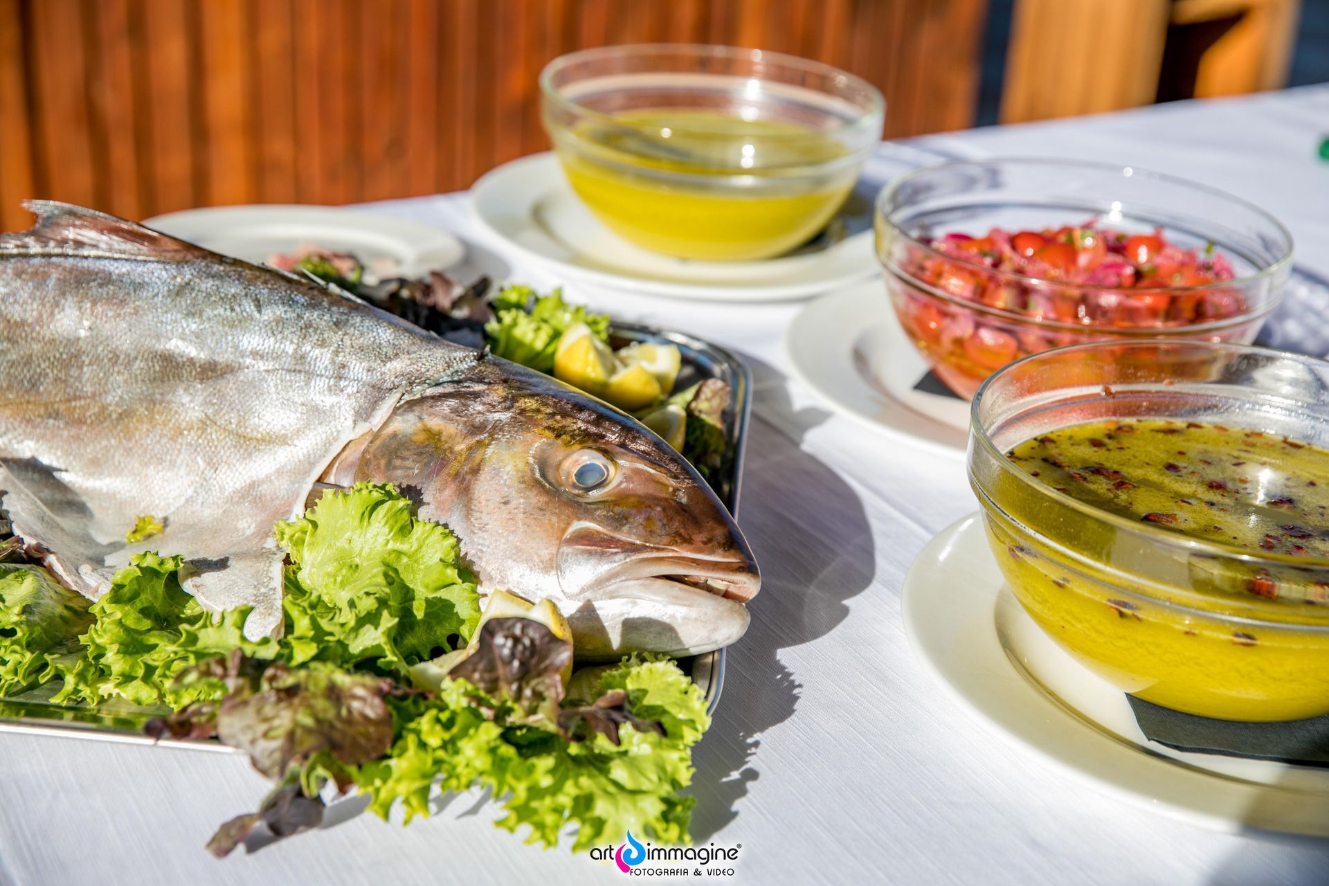 Pesce intero cotto su un piatto con insalata, circondato da piccole ciotole di salse su una tovaglia bianca.