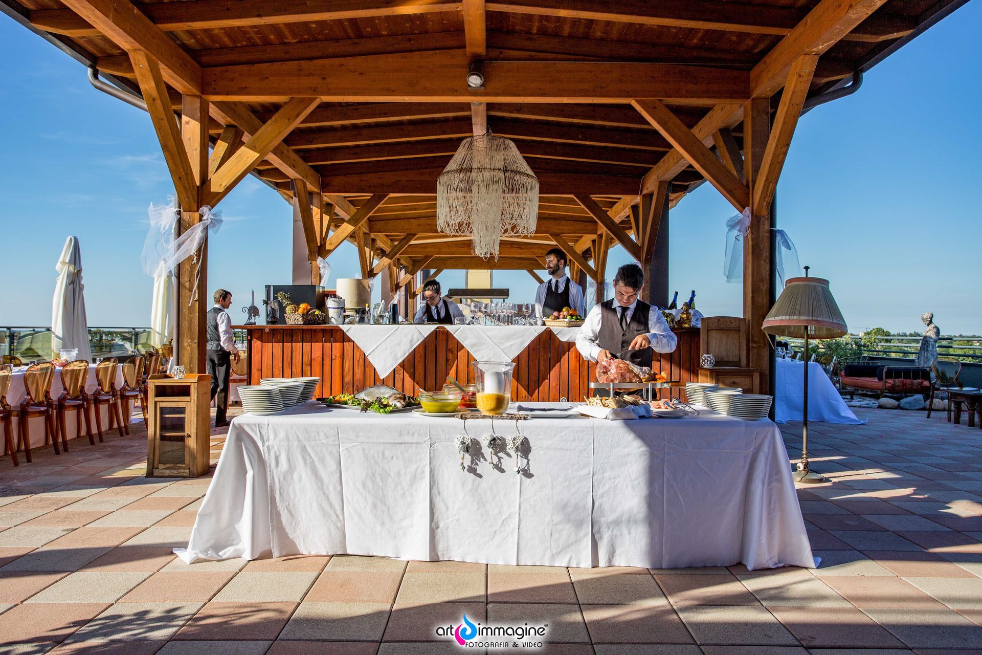 Allestimento per catering all'aperto sotto un pergolato in legno con cibo e camerieri.