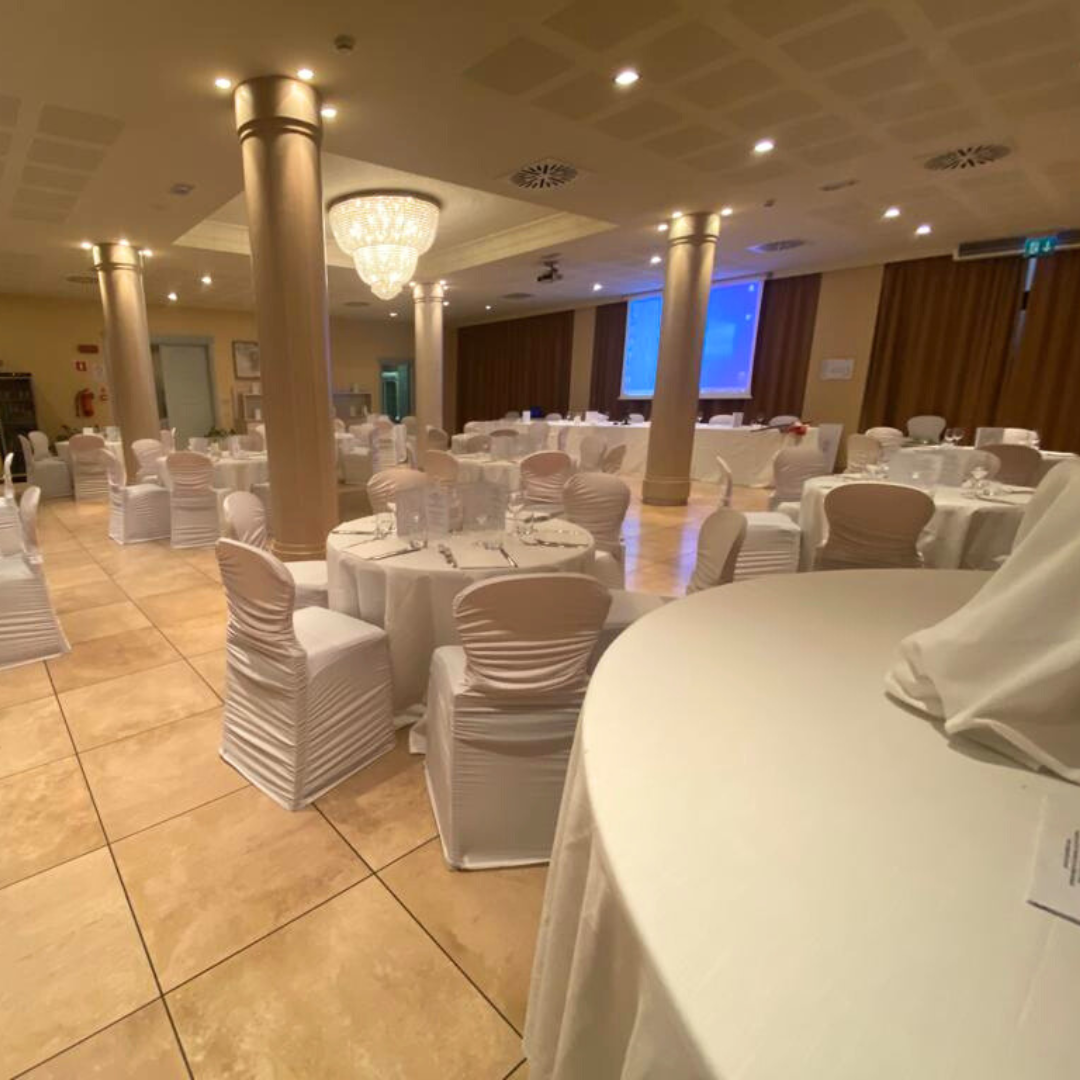 Elegante sala per banchetti con tavoli rotondi, sedie rivestite in tessuto bianco e un grande lampadario.
