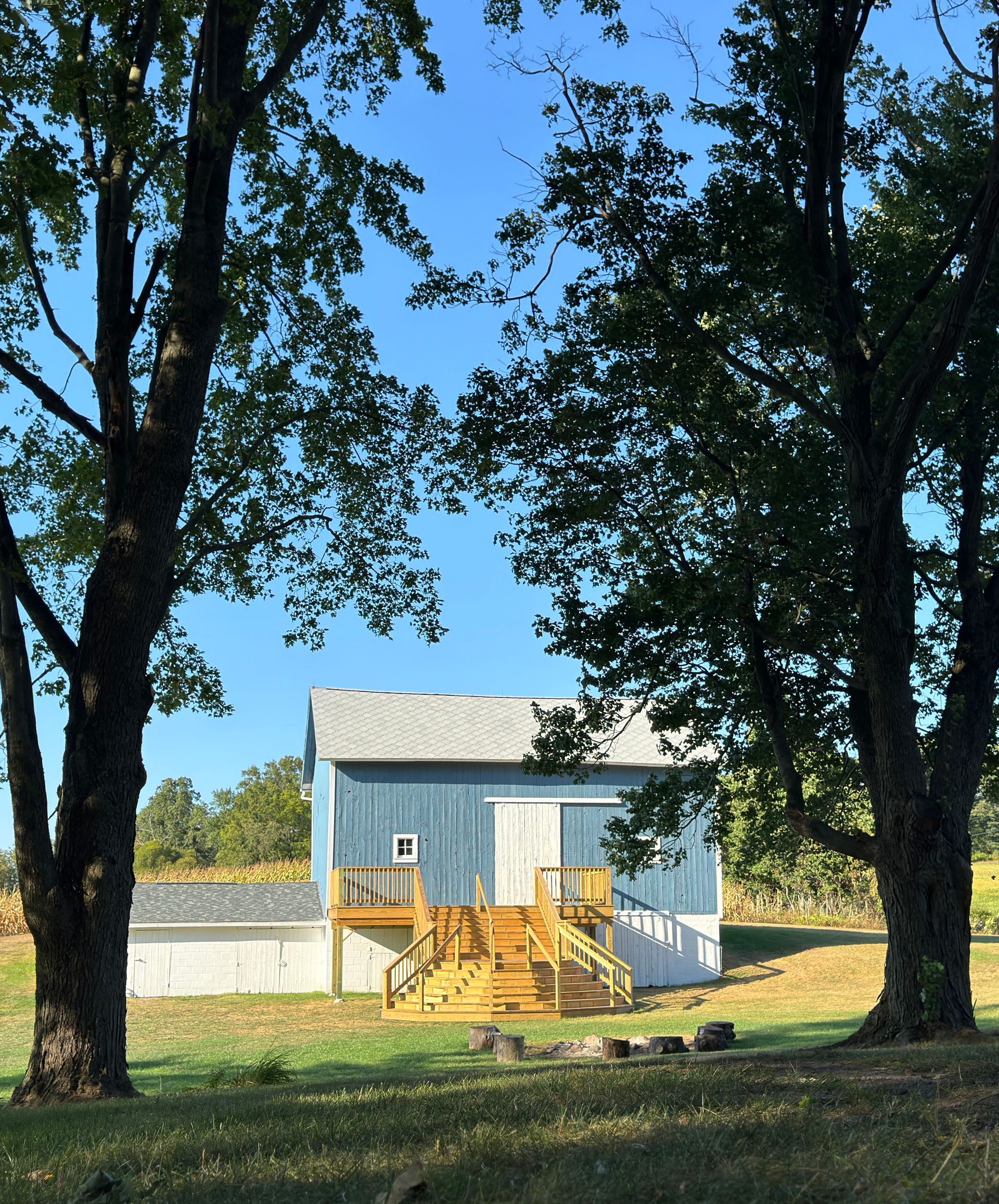 Blue wedding and event barn with wooden steps and a white outbuilding, framed by two trees, set 
in a grassy field under a blue sky.
