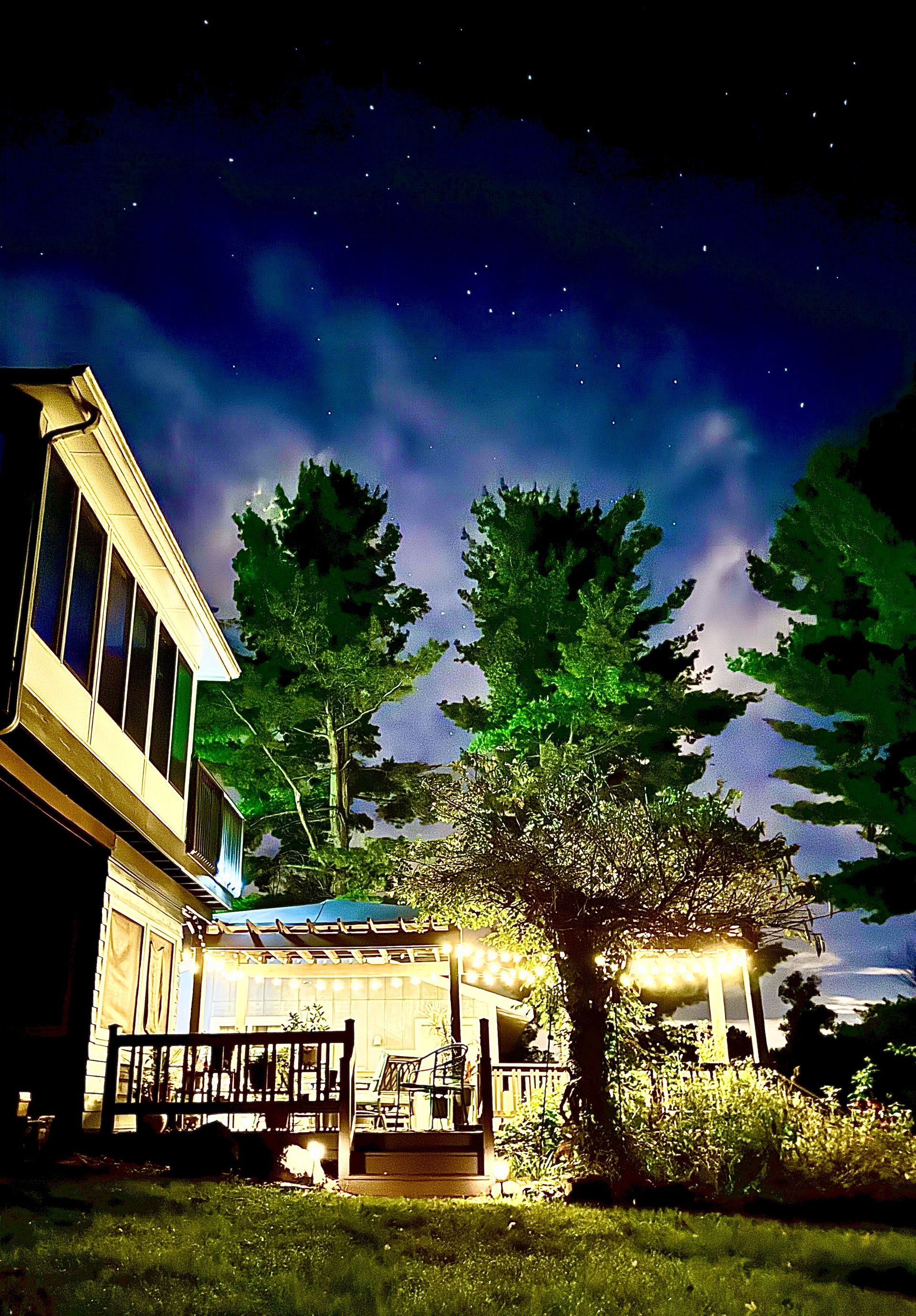 Nighttime scene: House with illuminated deck, trees, and starry sky.